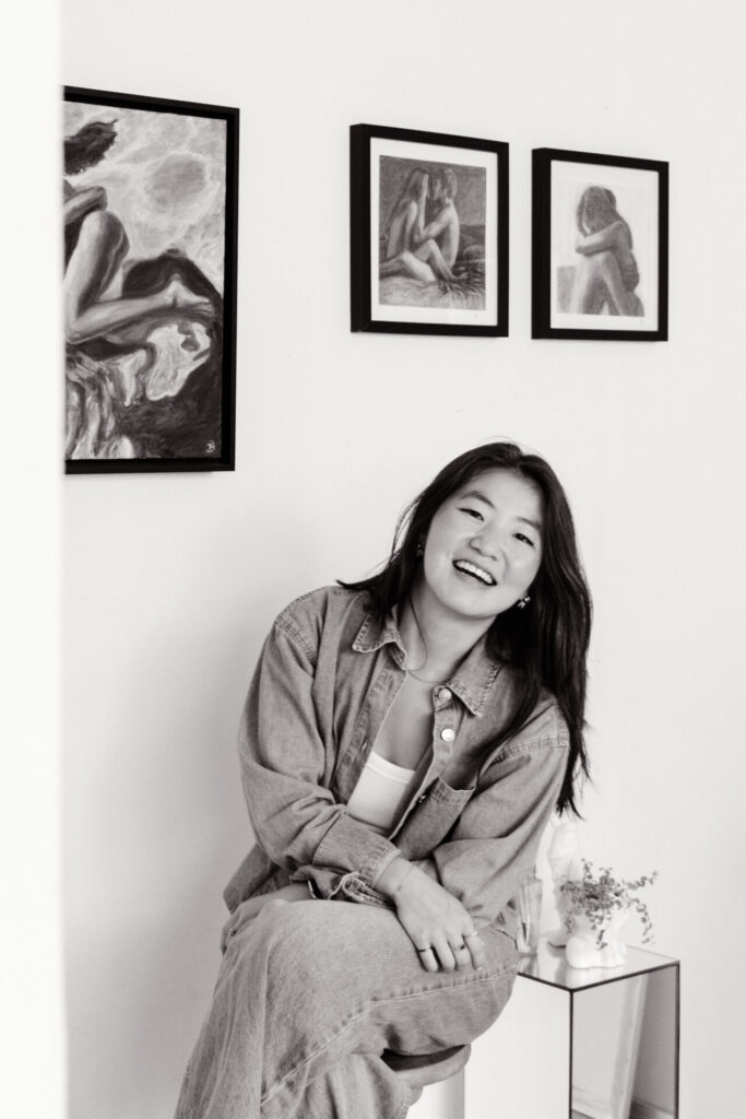Iris lachend in haar atelier, met ingelijste houtskooltekeningen aan de muur